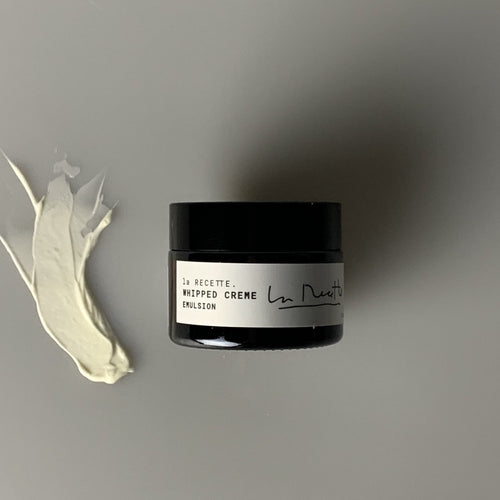 A dark glass jar with a white label that says Whipped Creme Emulsion in black elegant letters. on the left is a swipe of white face cream. 