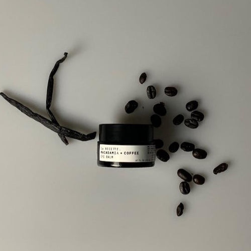 A dark amber low profile jar, with a white label which says la Recette. Macadamia + Coffee Eye Balm. coffee beans are scattered on the right vanilla bean pods are on the right.