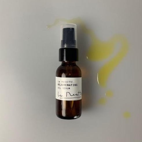 A small amber colored glass bottle with a white La Recette label saying Rejuvenating Oil Serum and a signature logo. There is a splash of gold  oil near the bottle. Image is shot from above. white background.