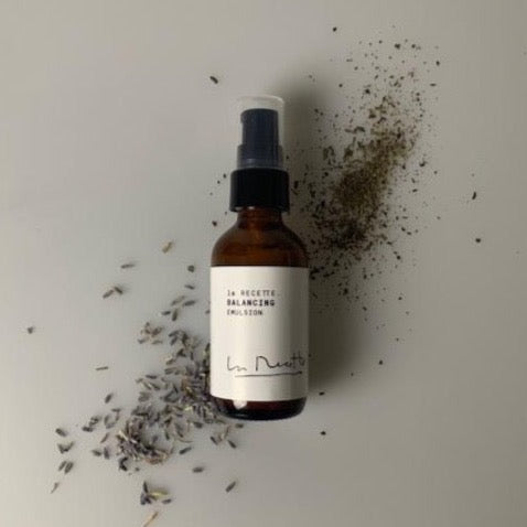 A photo of La Recette Skincare's Balancing Emulsion on a white background with a sprinkle of lavender and rosemary around it.